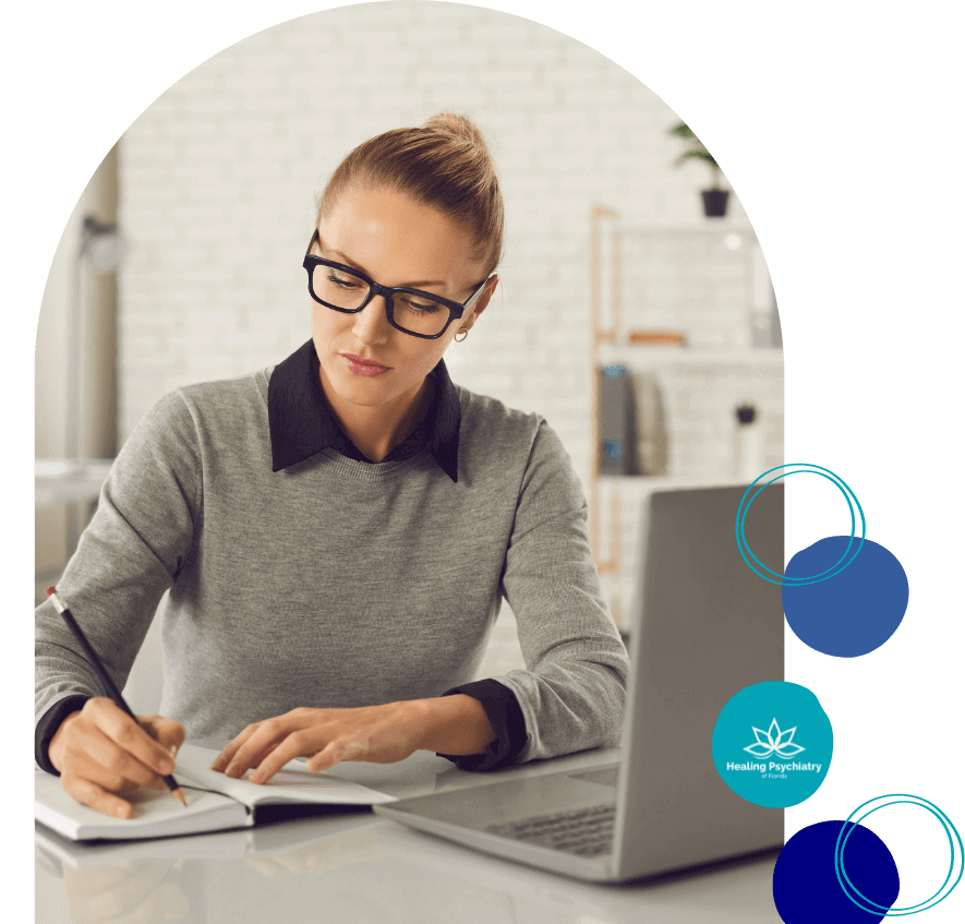 A focused professional woman is writing in a notebook at her desk with a laptop open in front of her