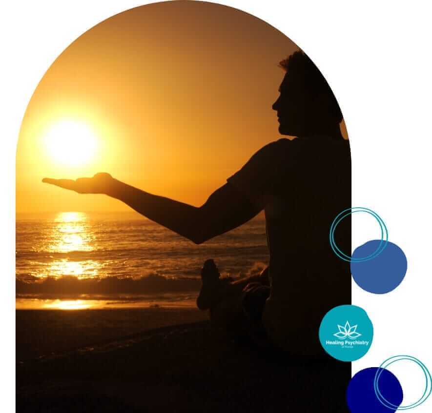 A silhouette of a person sitting cross-legged on the beach, facing a sunset over the ocean. The person is holding one hand up as if cradling the setting sun. The scene is tranquil, with the sun casting a golden glow over the water. On the right side of the image, there are blue and green circular logos with the text "Healing Psychiatry" and "Addiction Therapy in Lake Mary." The overall atmosphere is peaceful and reflective, suggesting a sense of healing and calm.