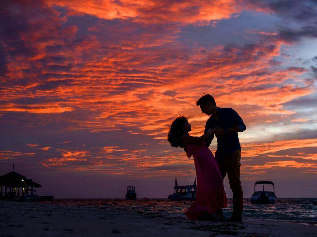 A couple dances on the beach at sunset with vibrant orange and red clouds in the sky, silhouetted against the ocean and boats in the background