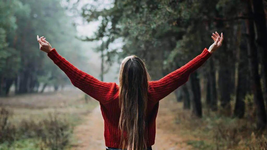 A woman with long hair stands in a forest clearing, arms raised in the air, wearing a red sweater, embracing nature