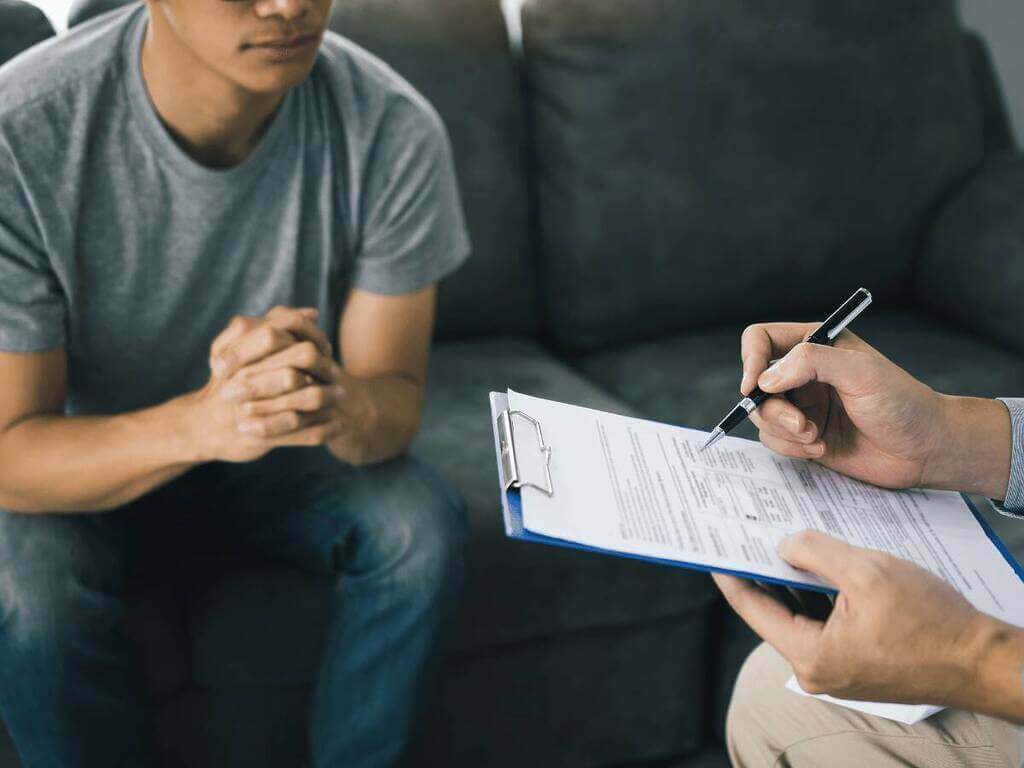 A patient sitting on a couch, hands clasped together, while a professional holds a clipboard and pen, seemingly discussing or assessing the situation, differences between Anxiety and PTSD.