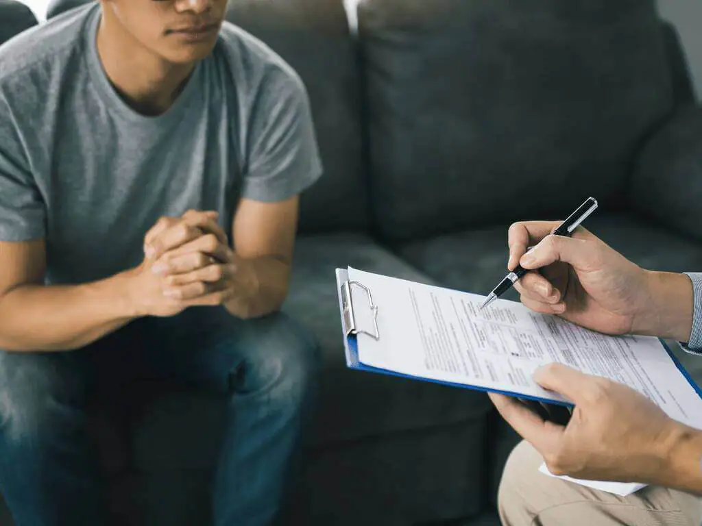 A patient sitting on a couch, hands clasped together, while a professional holds a clipboard and pen, seemingly discussing or assessing the situation, differences between Anxiety and PTSD.