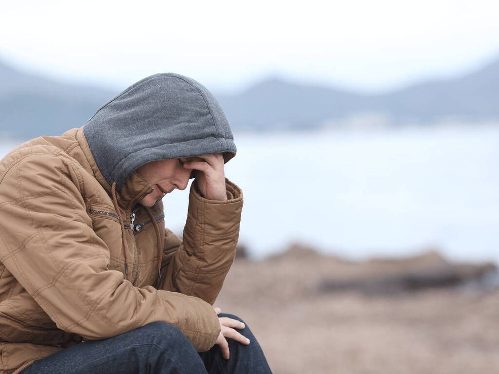 A person wearing a hooded jacket sitting alone outdoors, looking down in deep thought, differences between Anxiety and PTSD
