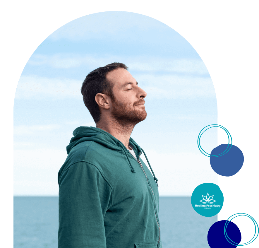 A man in a green hoodie stands peacefully by the ocean, taking deep breaths with his eyes closed. Symbolizing calm and recovery, this image reflects the journey of healing through opioid addiction treatment at Healing Psychiatry of Florida.