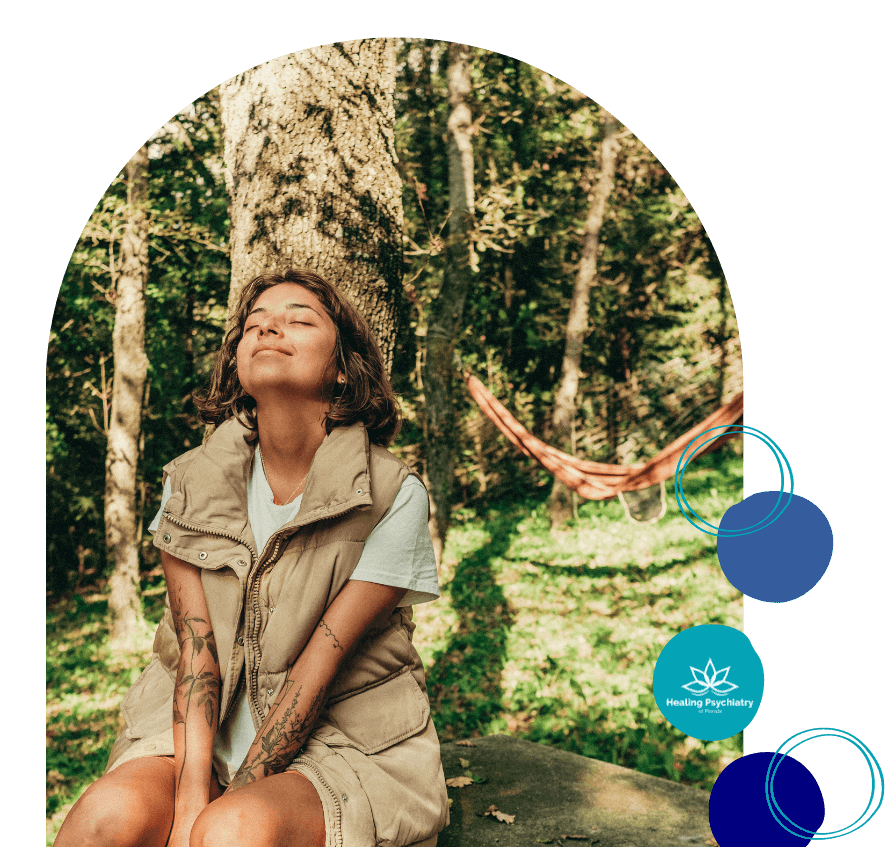 A woman with tattoos and a relaxed expression sits outdoors in a forested area, basking in the sunlight. This image represents the connection to nature and inner peace that is part of the holistic approach to opioid addiction treatment at Healing Psychiatry of Florida.