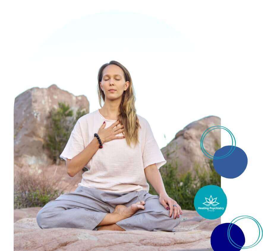 A woman sits cross-legged on a rocky surface, practicing mindfulness with her hand on her chest. This serene setting emphasizes the importance of meditation and emotional healing in opioid addiction recovery at Healing Psychiatry of Florida.