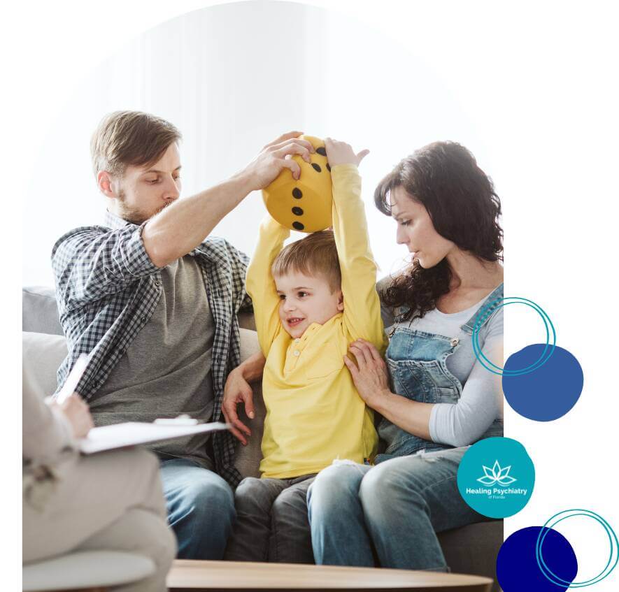 adhd kids (1) A child is playfully engaged with his parents during a therapy session, with one parent holding a yellow toy above the child’s head while the other parent supports him, as a therapist observes the interaction.