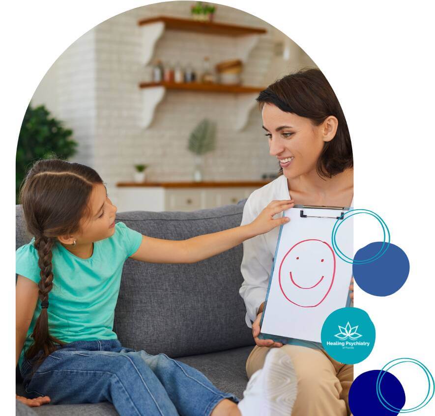 children therapy (4) A therapist and a young girl sitting on a couch, with the therapist holding a clipboard with a drawing of a smiley face while the girl points at it.