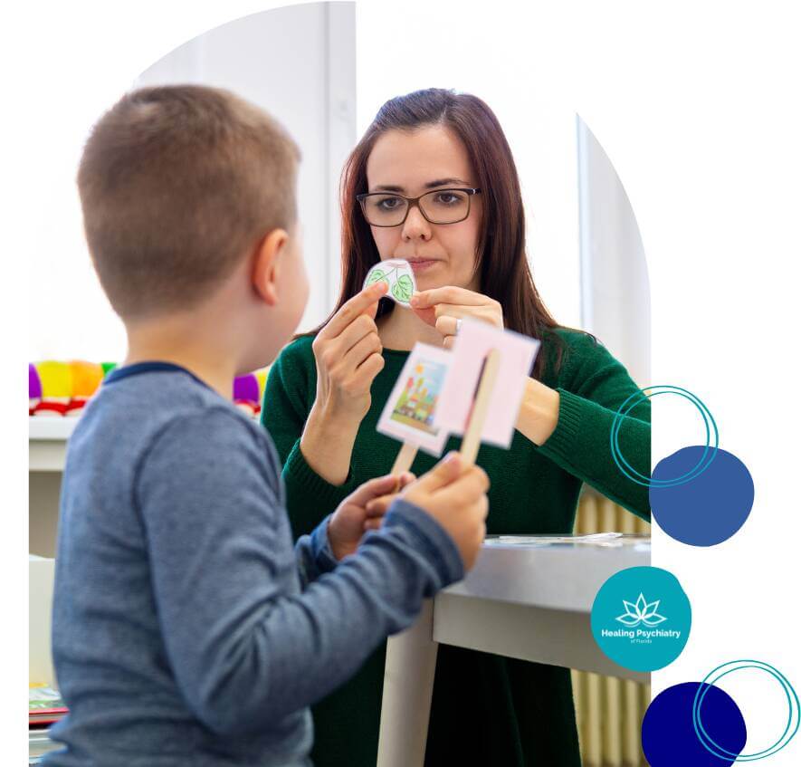 children therapy (5) A therapist and a young boy are sitting at a table during a therapy session. The therapist is holding up a card with a leaf drawing while the boy holds a card with a colorful picture, both focused on each other.