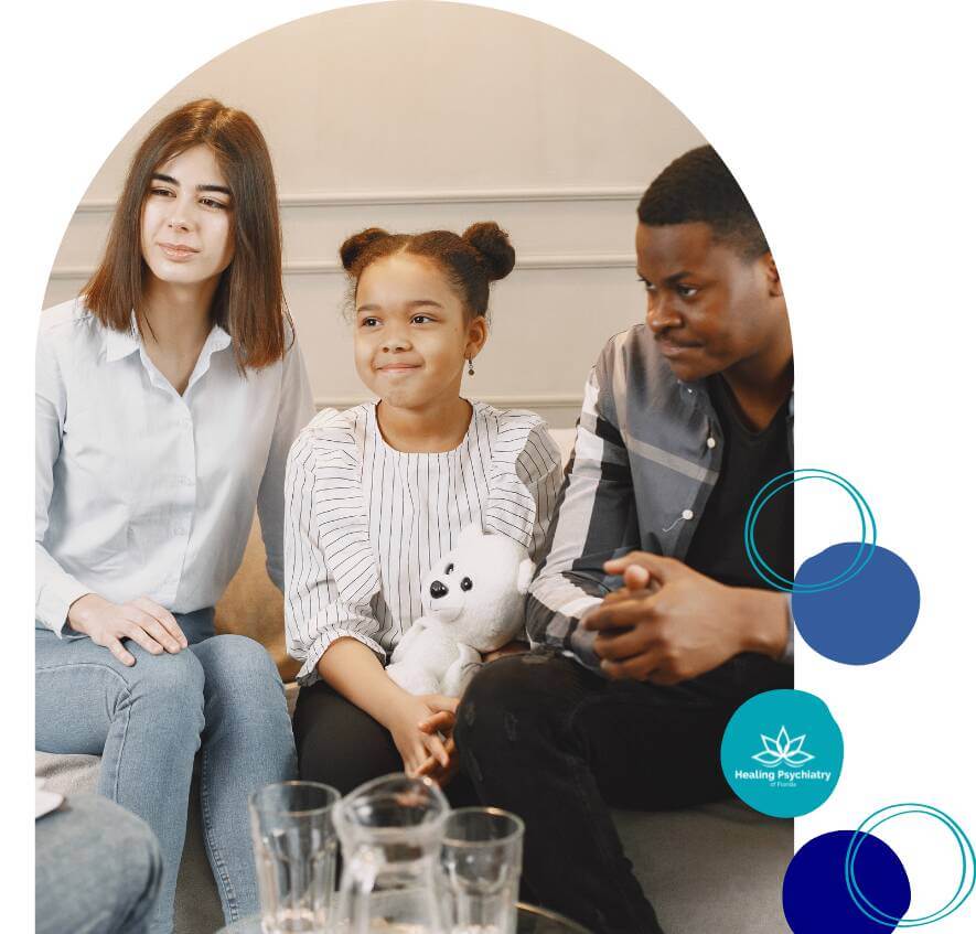 A child sits with two adults during a therapy session, smiling with a stuffed bear in her hands, while the adults provide support and guidance.