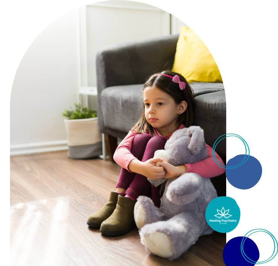 A young girl sits on the floor, hugging a teddy bear and looking down with a sad expression in a cozy, quiet room.