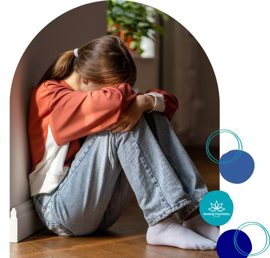 A teenage girl sits on the floor with her head resting on her knees, showing signs of sadness and distress in a quiet, peaceful room.