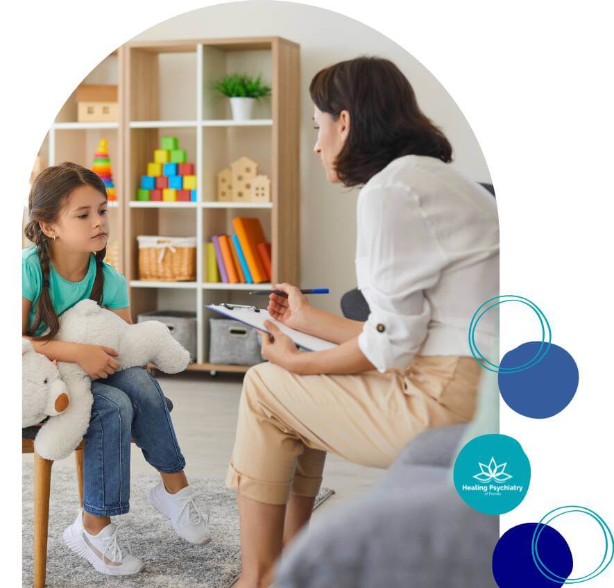 A young girl holds a teddy bear while sitting on a chair during a therapy session, listening attentively to a therapist who is taking notes. The room is bright and welcoming with colorful toys in the background.