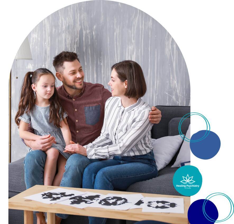 A family with a young daughter sits on a couch during a therapy session, with inkblot test papers on a table in front of them.