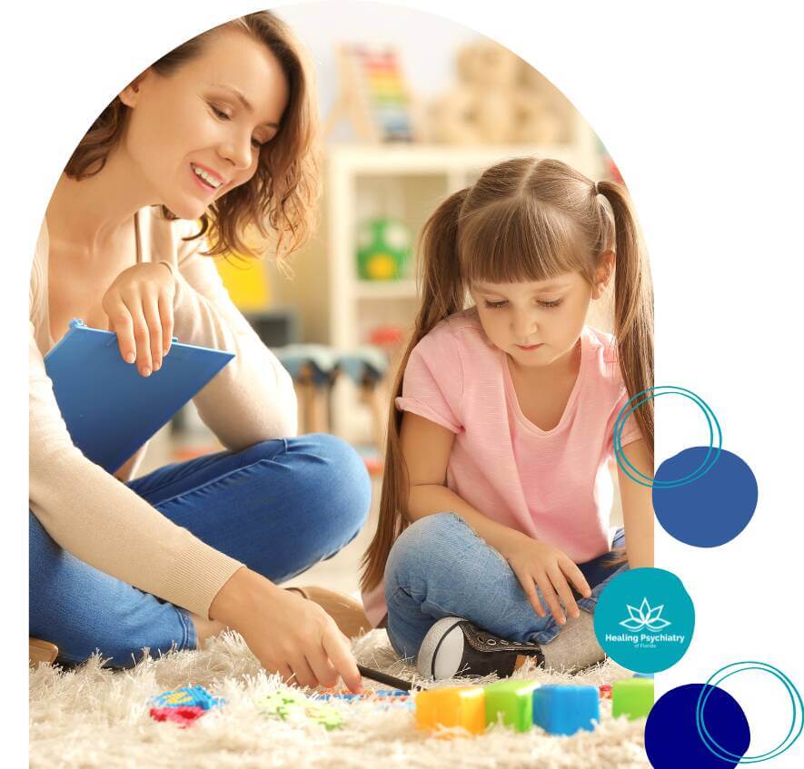 A young girl participates in a play therapy session with a smiling therapist, using colorful blocks on the floor.