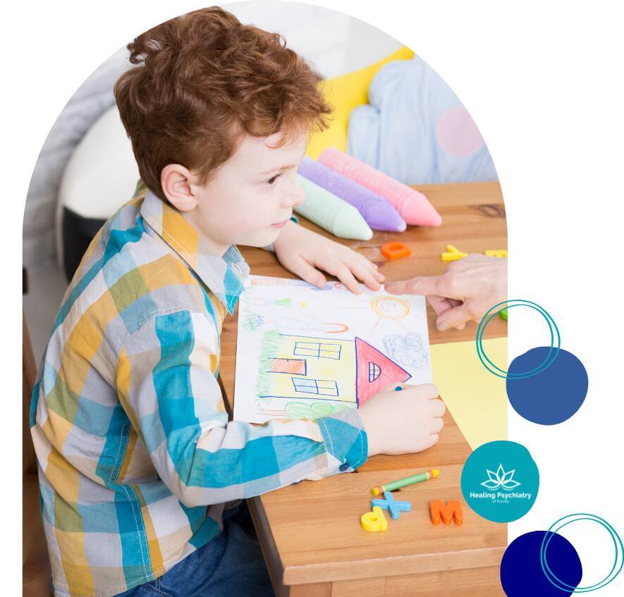 A young boy sits at a table during a therapy session, showing a drawing of a house to a therapist.