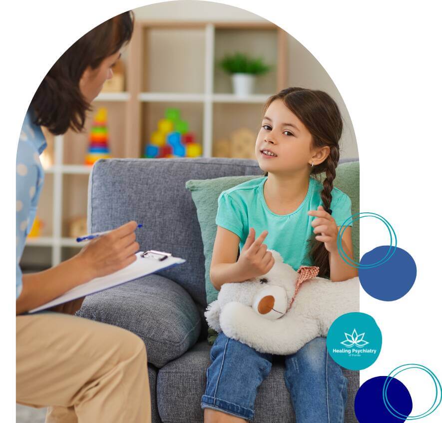 A young girl hugs a stuffed animal while talking to a therapist holding a clipboard during a therapy session.