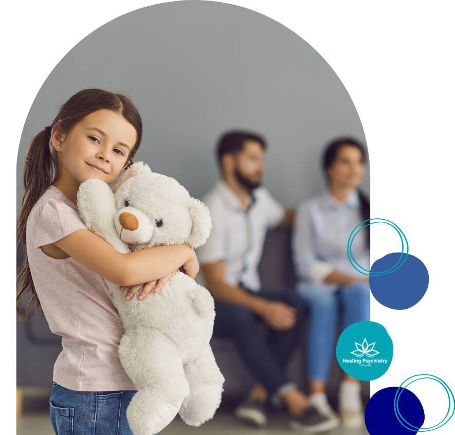 A young girl smiles while hugging a white teddy bear, standing in front of two adults seated in the background. 