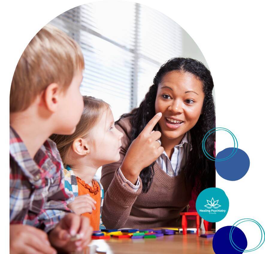 A therapist engages two children with facial cues during an emotional regulation session, using colorful tools at a table.
