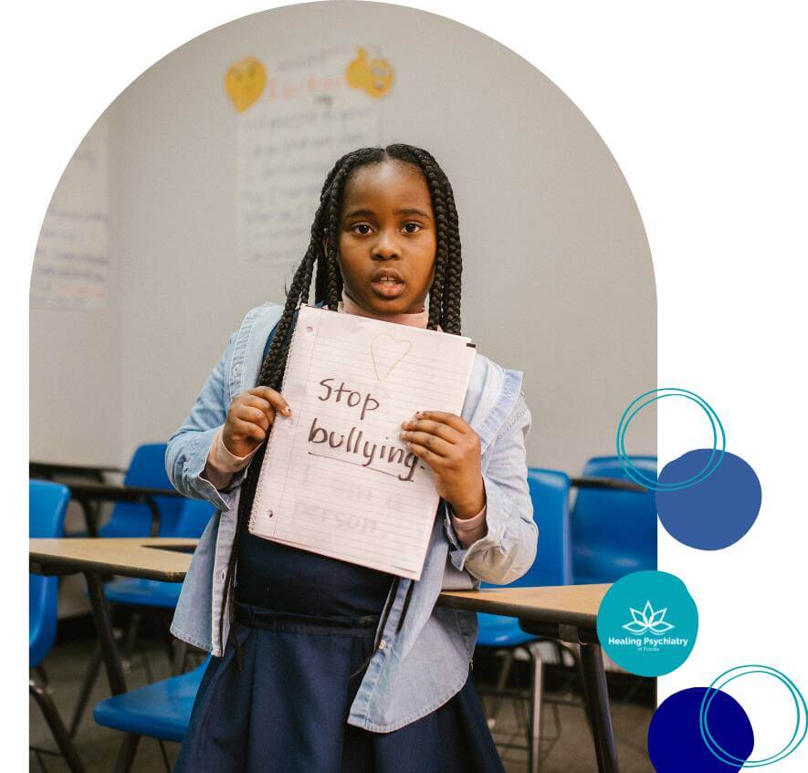 A young girl standing in a classroom holds up a notebook with the handwritten message “Stop bullying” and a heart drawn above it.