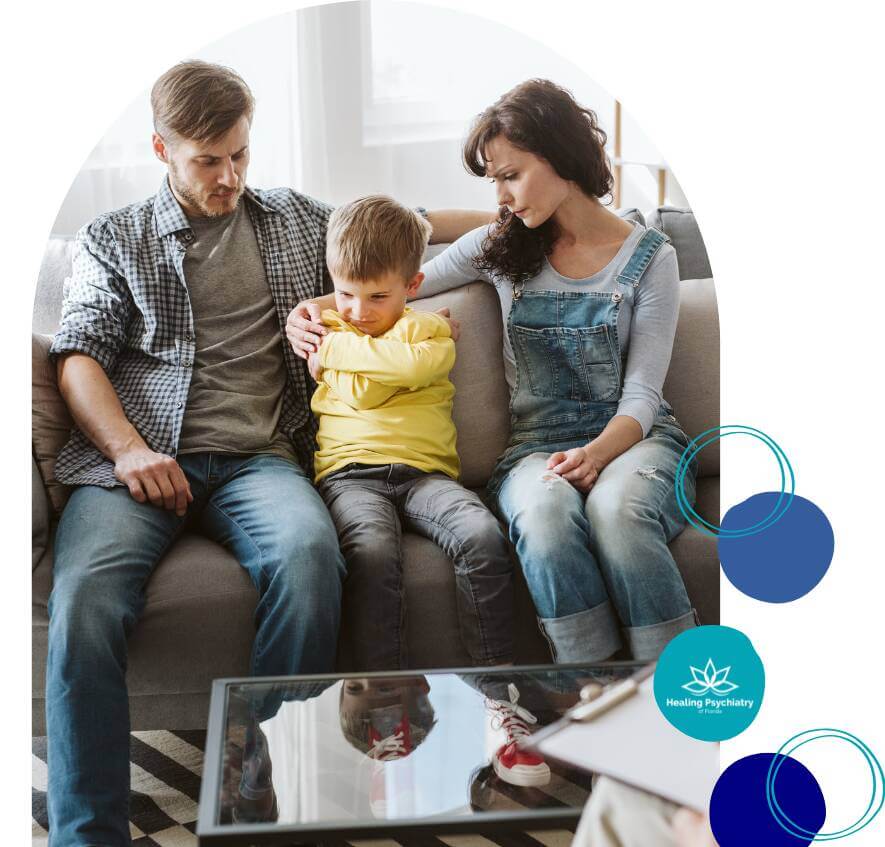 A young boy sits between his concerned parents on a couch, arms crossed and looking upset, during a family therapy session.