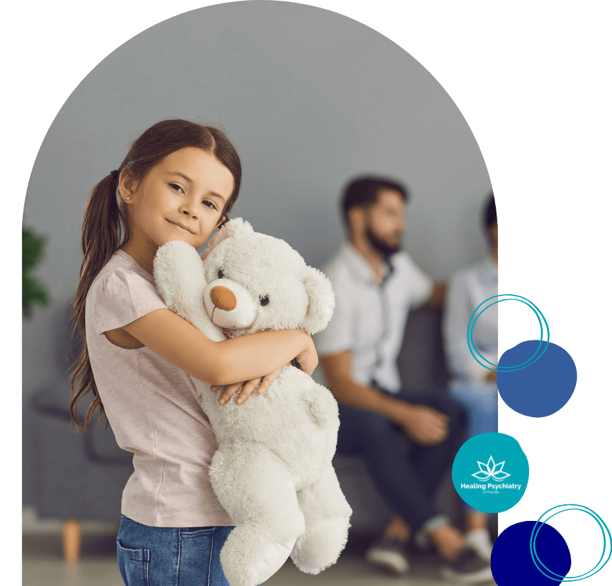 Young girl hugging a white teddy bear and smiling, with two blurred adults sitting in the background, representing children’s therapy and emotional support during divorce at Healing Psychiatry of Florida.