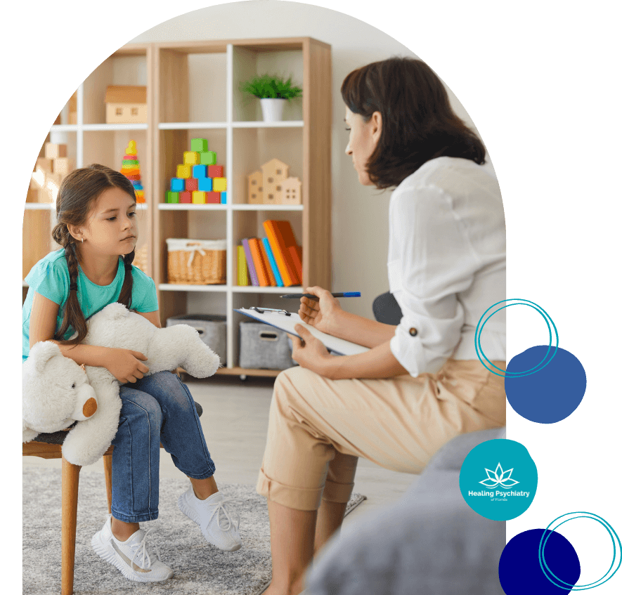 A young girl sits on a chair holding a white teddy bear, looking sad and thoughtful during a therapy session. A female therapist sits across from her with a clipboard and pen, listening attentively. They are in a child-friendly room with shelves of colorful toys and books. The image includes the Healing Psychiatry of Florida logo.<br />
