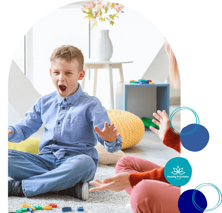 A young boy sits cross-legged on the floor with his mouth open and arms raised, appearing to shout or express frustration. He wears a blue button-up shirt and jeans. An adult, partially visible, is seated across from him, gesturing with open hands. Toys are scattered on the carpet, and the room has a calm, child-friendly setting. The image includes the logo for "Healing Psychiatry of Florida."