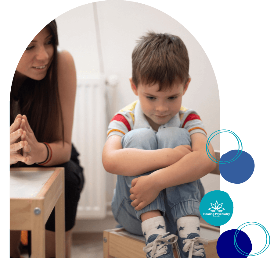 A young boy sits on a wooden stool with his knees pulled to his chest, looking down with a sad or withdrawn expression. A woman beside him, possibly a therapist or caregiver, watches him with a calm and attentive posture. The image includes a logo that reads "Healing Psychiatry of Florida.