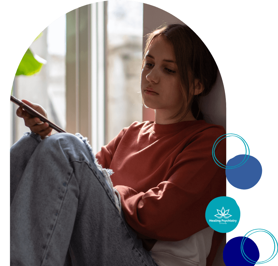 A teenage girl sits by a window holding her phone, looking thoughtful and distant.