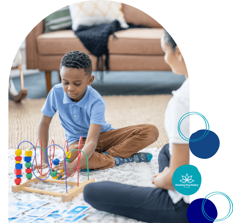 A young boy sits on a carpeted floor playing with a colorful bead maze toy during a therapy session. An adult, possibly a therapist, sits nearby observing and engaging with him. The setting appears calm and home-like, with a couch and pillows in the background. The image includes a logo that reads "Healing Psychiatry of Florida.