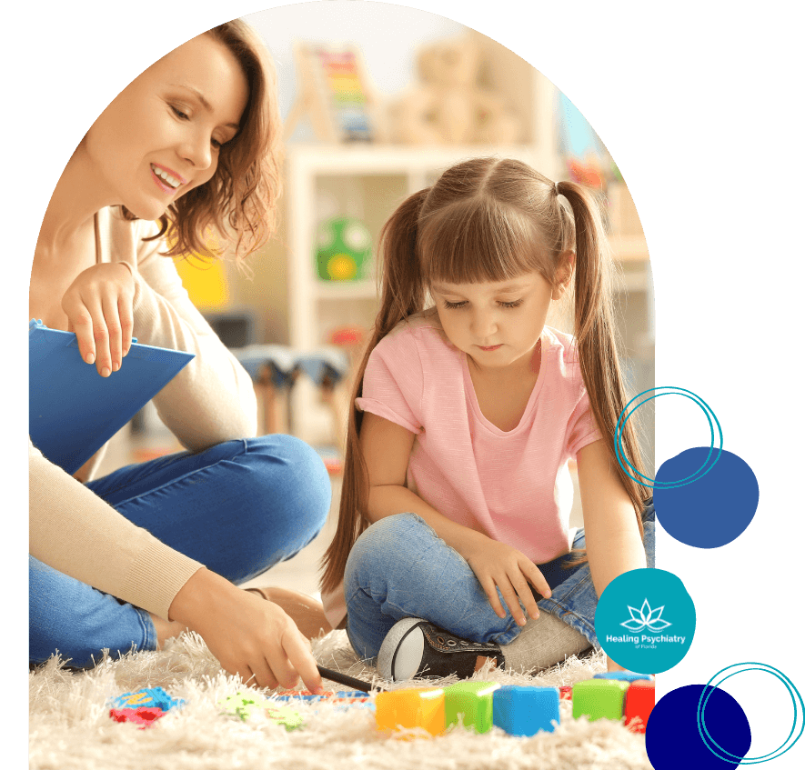 A young girl sits on a soft rug playing with colorful building blocks while a female therapist observes and takes notes with a clipboard. They are in a brightly lit room with toys and shelves in the background. The image includes a logo that reads "Healing Psychiatry of Florida.