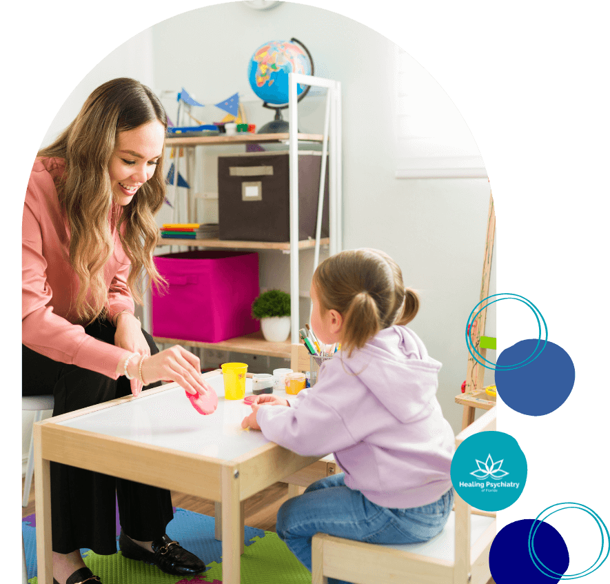 A young girl sits at a small table across from a female therapist in a brightly colored therapy room. The therapist, smiling, uses playdough during their session, encouraging interaction. The room contains educational toys, shelves, and a globe. A logo reading "Healing Psychiatry of Florida" appears in the image.