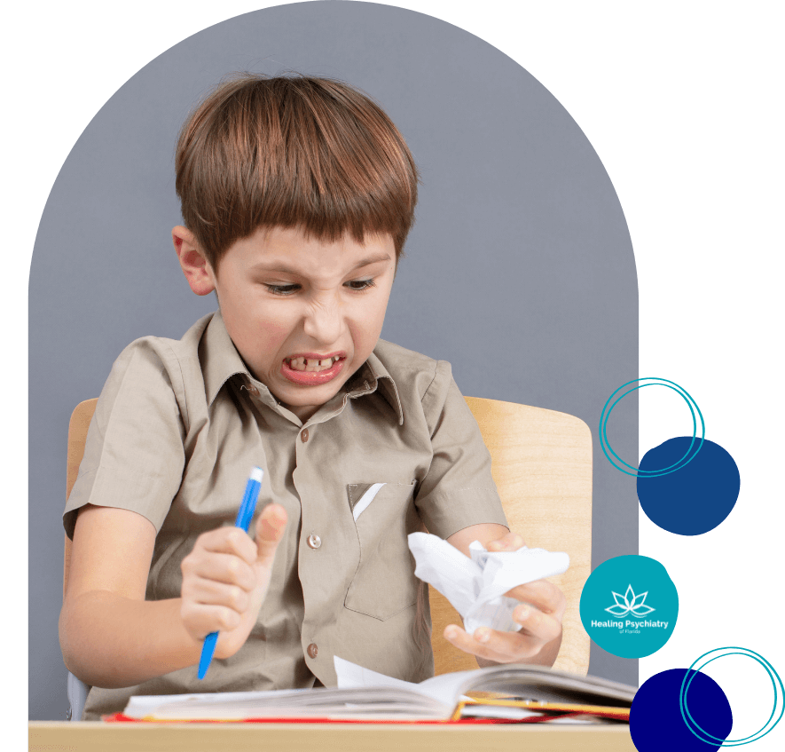 anger A frustrated young boy sits at a desk gripping a pen and crumpled paper, showing signs of anger and stress while doing schoolwork. The “Healing Psychiatry of Florida” logo appears in the lower corner.