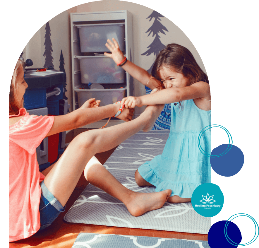 anger1 Two young girls sit on play mats in a room, engaged in a physical struggle over a toy with tense expressions. The scene suggests sibling conflict or behavioral challenges. The "Healing Psychiatry of Florida" logo appears in the corner.<br />