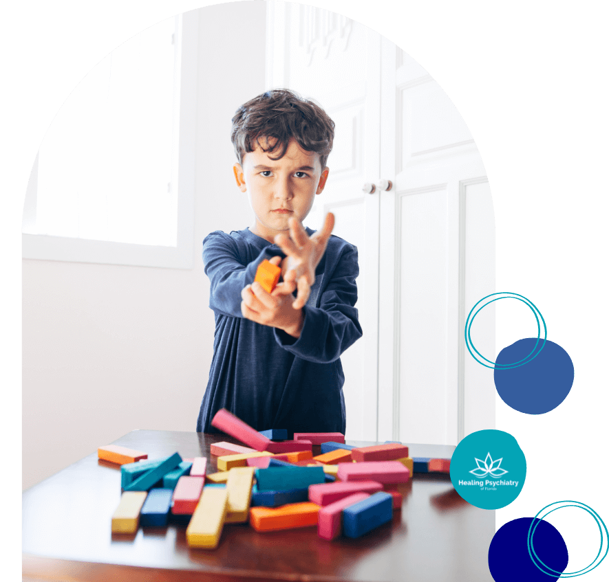 anger2 A young boy stands at a table with colorful wooden blocks scattered in front of him, throwing one block with a frustrated expression. The scene suggests emotional dysregulation or impulsive behavior. The “Healing Psychiatry of Florida” logo is visible on the right side.