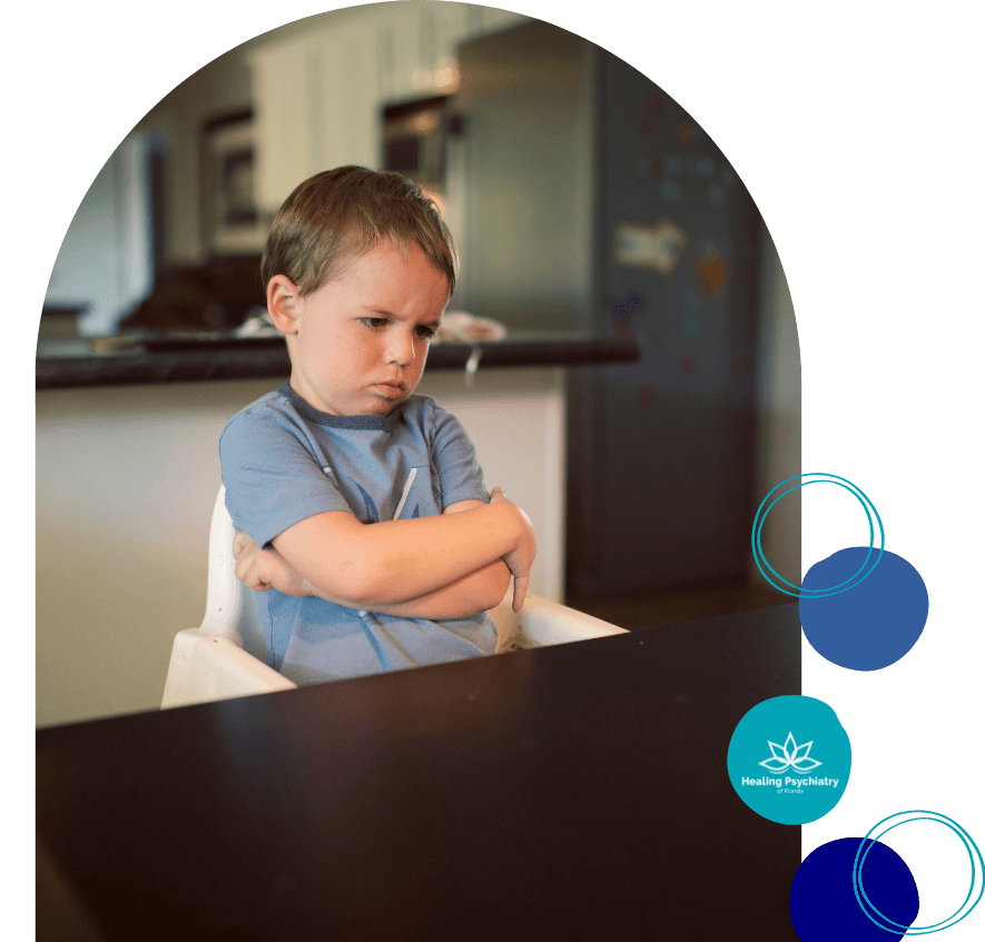 anger3 A young boy sits in a high chair with his arms crossed and an upset expression on his face, looking down at the table. The kitchen setting in the background suggests a home environment. The image conveys feelings of frustration or defiance. The “Healing Psychiatry of Florida” logo appears on the right side.
