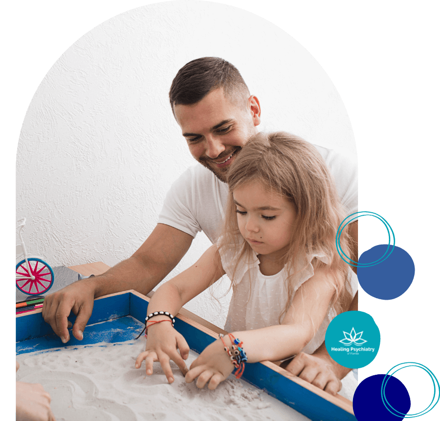 sand therapy1 A young girl focuses intently while playing in a sand tray, as a smiling adult male—possibly a therapist or parent—watches supportively. The image includes the Healing Psychiatry of Florida logo, suggesting a therapeutic setting.