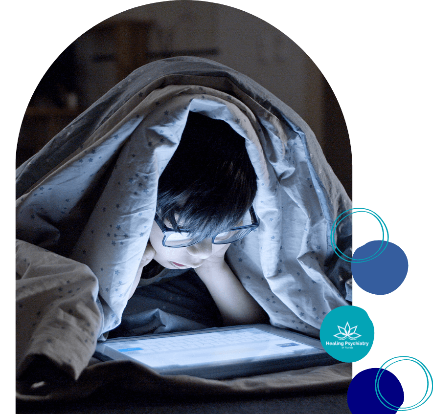 A young boy uses a tablet under a blanket at night, with the screen lighting up his face.